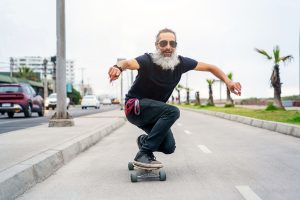 A person with long hair and a beard skateboards down a road, balancing skillfully. They wear sunglasses, a black t-shirt, and jeans, with a flannel shirt tied at the waist. Cars and palm trees line the background under a cloudy sky.