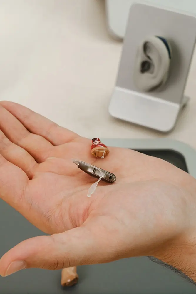 A person holds two types of hearing aids in their palm. In the background, there is an ear model on a display stand.